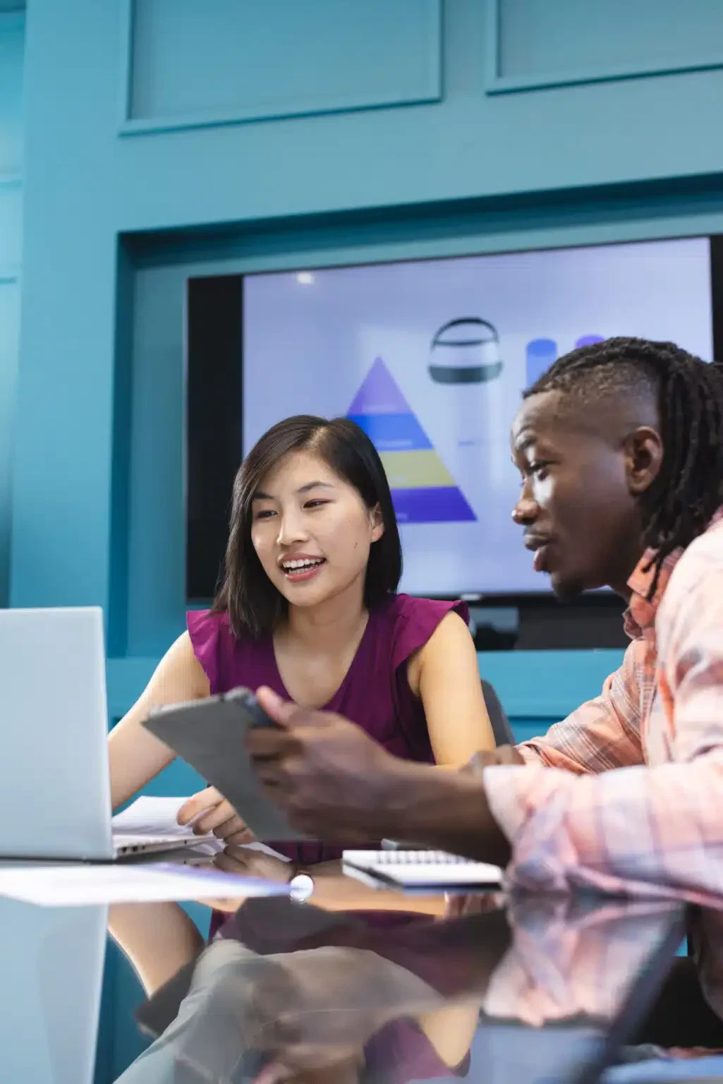 African American man and Asian woman are looking at laptop in a modern business office. Both are young, he wearing orange shirt, she in purple top, unaltered.