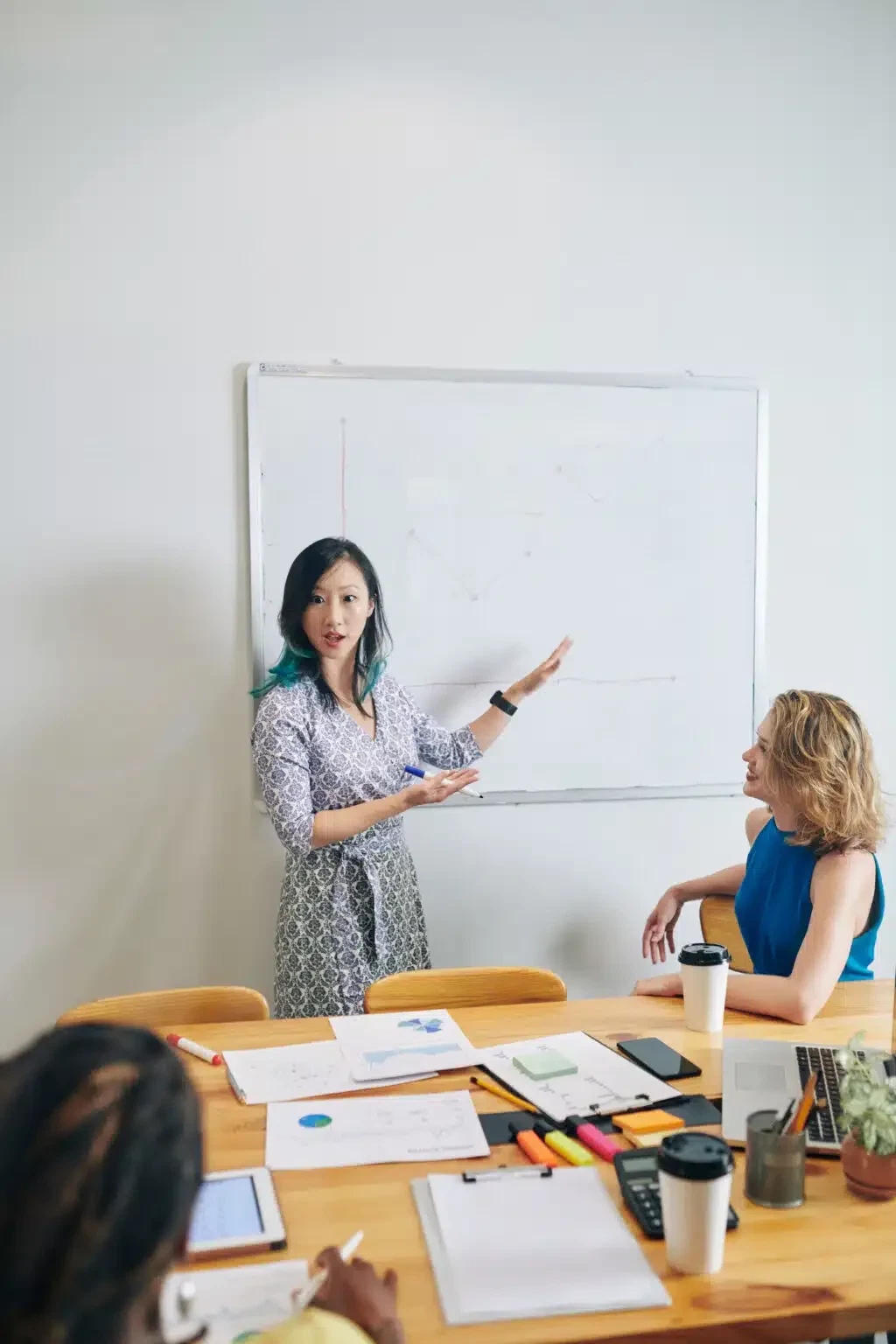 Young Chinese businesswoman showing chart on whiteboard when talking to her collagues at meeting during coronavirus crisis